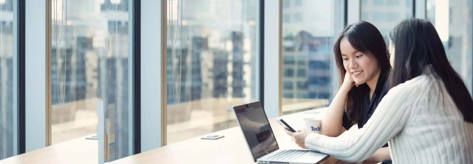 Two women working on a laptop by large windows with a city view.