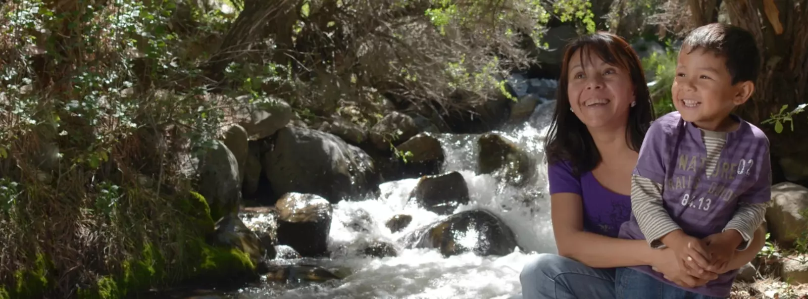 A woman and child smiling by a forest stream.