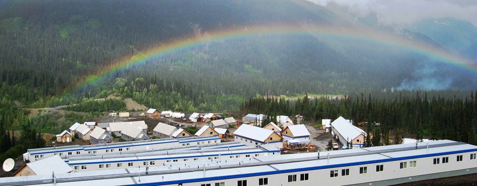 A rainbow arcs over a small village with white-roofed buildings surrounded by forested mountains.