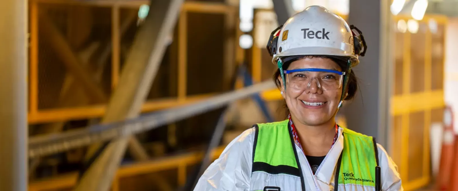 Smiling worker in safety gear and helmet with 'Teck' logo in an industrial setting.