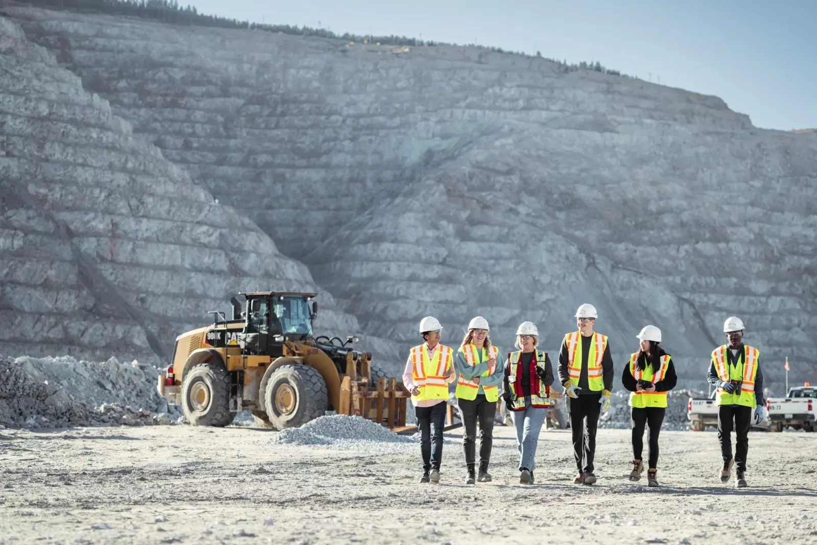 Six workers in safety gear walk through a quarry, with excavator in the background.