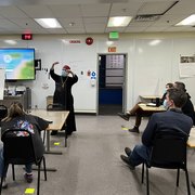 Person presenting to seated individuals in a classroom with a wall-mounted screen.