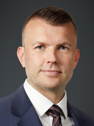 Man in a suit with a serious expression against a dark background.