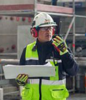 Construction worker in safety gear talking on a radio, holding plans at a work site.