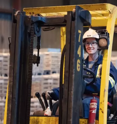 Smiling worker in safety gear operating a yellow forklift inside an industrial warehouse.