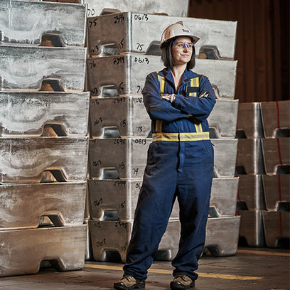 Worker in blue coveralls and helmet stands in front of stacked metal ingots in an industrial setting.