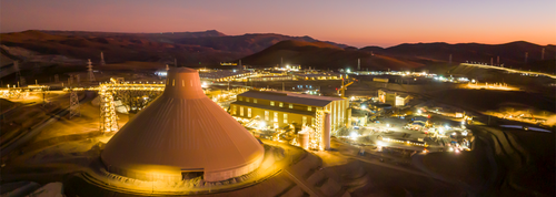 Illuminated industrial complex at dusk surrounded by rolling hills and distant mountains.