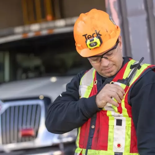 Worker in high-visibility vest and hard hat using walkie-talkie near large truck.