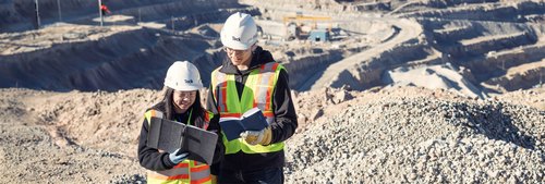 Two workers in safety gear reviewing plans at a mining site.