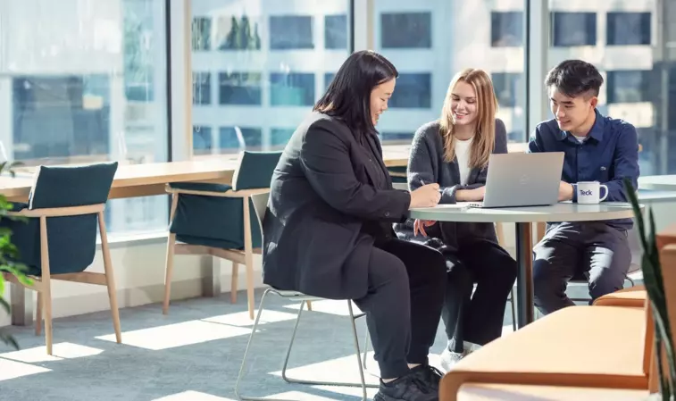 Three people collaborating at a table with a laptop in a modern office setting.