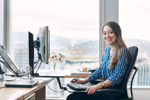 Smiling person working at a desk with a computer in a bright office setting.