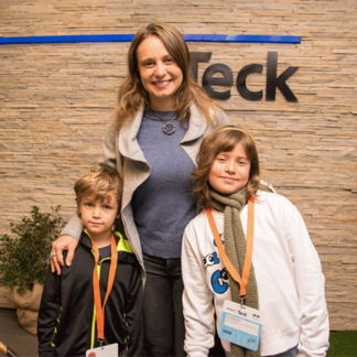 A woman with two children stands in front of a wall with a 'Teck' sign, all smiling at the camera.