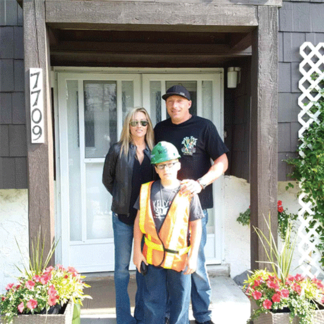 A family of three posing in front of a house with the number 7709 displayed on the door frame.