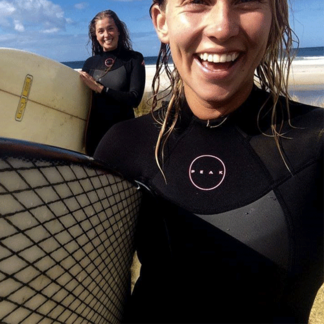 Two women in wetsuits holding surfboards, smiling by the beach.