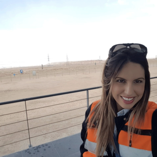 Smiling woman in an orange safety vest outdoors with a desert landscape in the background.