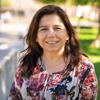 Woman with long dark hair and floral blouse smiling outdoors on a sunny day.