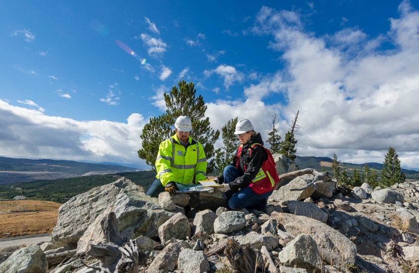 Two workers in safety gear, including hard hats and jackets, examining documents on a rocky landscape with a scenic mountainous background