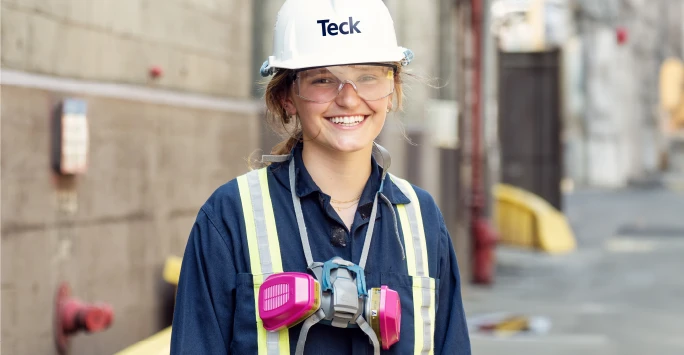 Smiling worker in safety gear and helmet, standing outdoors near an industrial building.