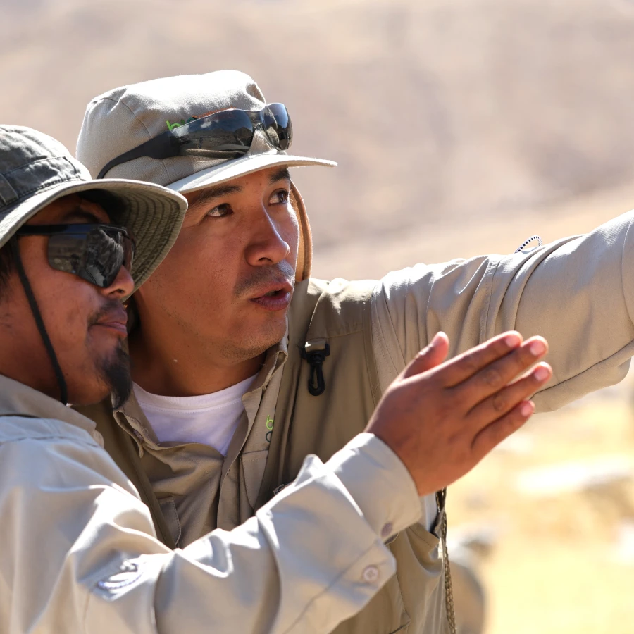 Two anglers in sun hats collaborate, pointing toward an unseen object in a desert landscape.