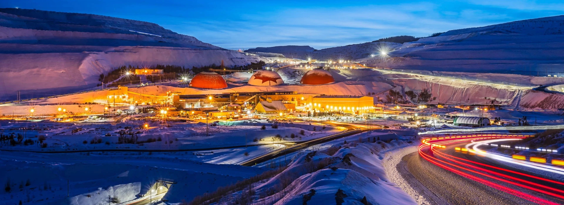 Illuminated mining facility at dusk surrounded by snowy landscape with trails of light from passing cars.