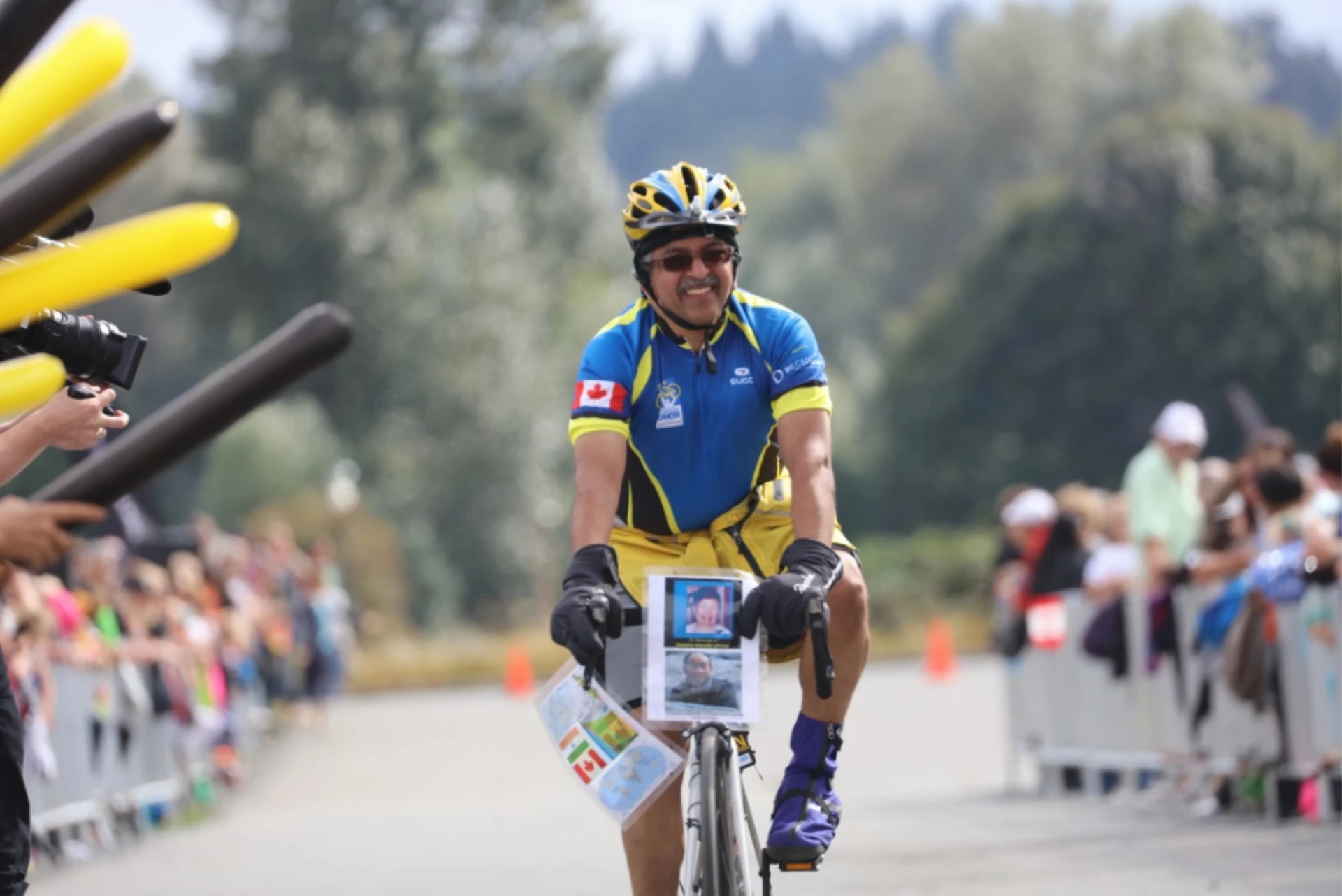 Cyclist in a race, wearing a blue and yellow helmet, smiling, with cheering crowd in the background.